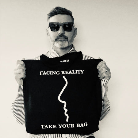 Schwarze TAKE YOUR BAG mit weißem FACING REALITY Design aus 100% Bio-Baumwolle, NEUTRAL® und FAIRTRADE® getragen von einem Man mit der schwarzen Sonnenbrille.