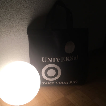 Schwarze TAKE YOUR BAG mit weissen UNIVERSAL  Design aus 100% Bio-Baumwolle, NEUTRAL® und FAIRTRADE® zertifiziert hinter der Lampe.