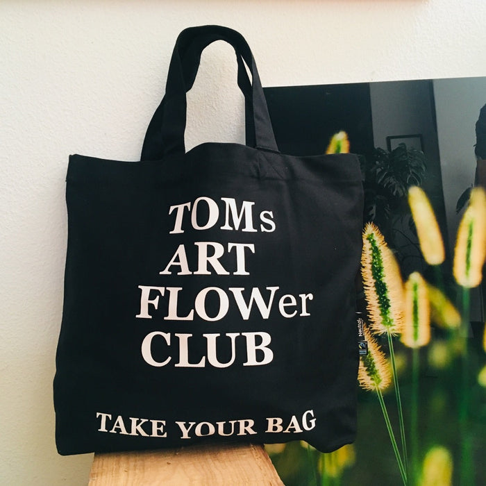Schwarze TAKE YOUR BAG mit weißem TOMs FLOWer CLUB Design aus 100% Bio-Baumwolle, NEUTRAL® und FAIRTRADE® zertifiziert vor dem Bild.