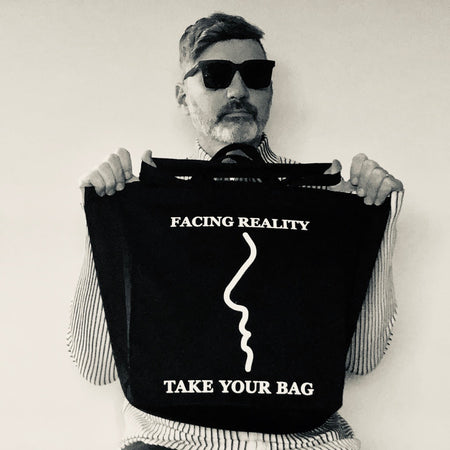 Schwarze TAKE YOUR BAG aus 100% Bio-Baumwolle, NEUTRAL®- und FAIRTRADE®-zertifiziert mit weißem FACING REALITY-Design, getragen von einem Mann mit der schwarzen Sonnenbrille.