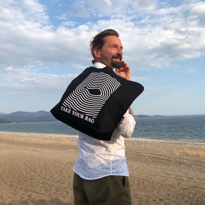 Schwarze TAKE YOUR BAG mit weißem FISH-Motiv aus 100% Bio-Baumwolle, NEUTRAL® und FAIRTRADE® zertifiziert getragen von einem Mann auf dem Strand.