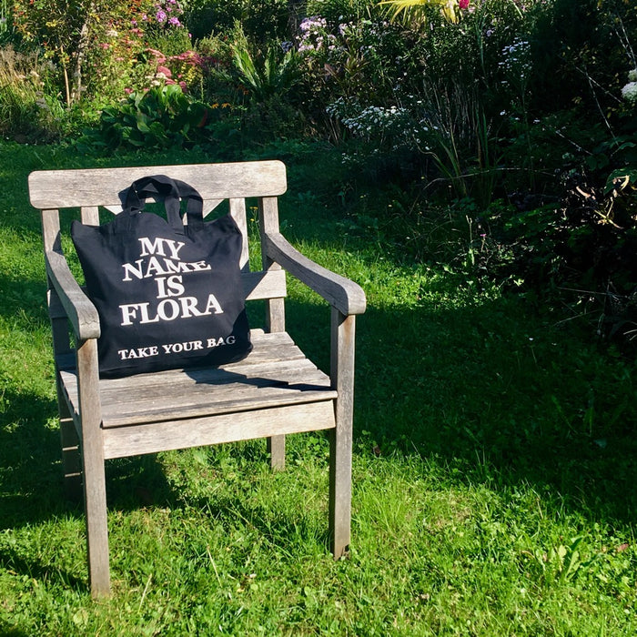 Schwarze TAKE YOUR BAG mit weißem MY NAME IS FLORA Motiv aus 100% Bio-Baumwolle, NEUTRAL® und FAIRTRADE®  auf der Gartenbank.