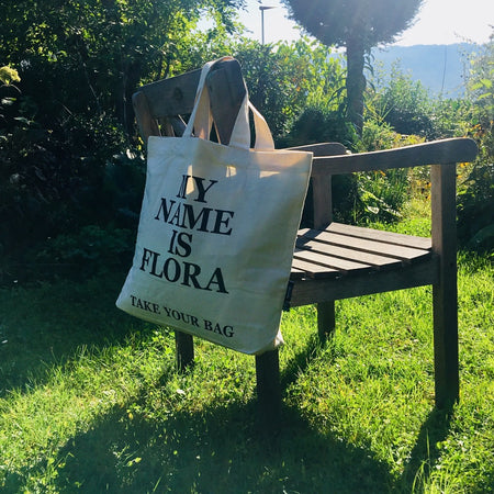 Beige TAKE YOUR BAG mit schwarzem MY NAME IS FLORA Design aus 100% Bio-Baumwolle, NEUTRAL® und FAIRTRADE® zertifiziert auf dem Gartenstuhl im Garten.