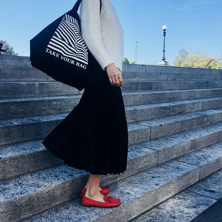 Schwarze TAKE YOUR BAG aus 100% Bio-Baumwolle, NEUTRAL®- und FAIRTRADE®-zertifiziert mit weißem LINEAR-Design, getragen von einer Frau mit roten Schuhen auf der Treppe.
