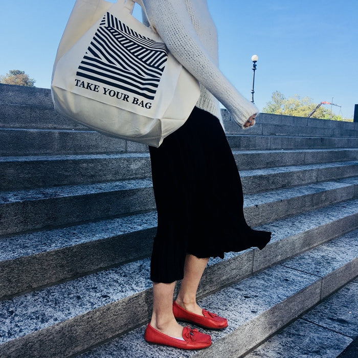 Beige TAKE YOUR BAG aus 100% Bio-Baumwolle, NEUTRAL® und FAIRTRADE® zertifiziert mit schwarzem FACING REALITY Design, getragen von einer Frau mit roten Schuhen auf der Treppe..
