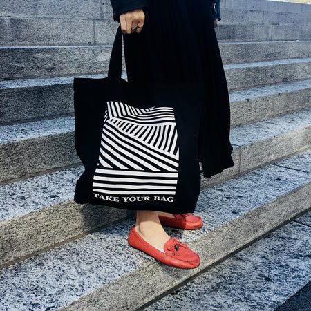 Schwarze TAKE YOUR BAG mit weißem LINEAR Design aus 100% Bio-Baumwolle, NEUTRAL® und FAIRTRADE® zertifiziert getragen von einer Frau mit roten Schuhen auf der Treppe.