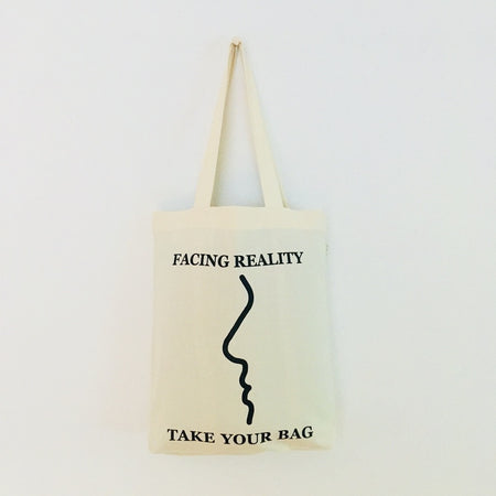A beige cotton tote bag with a black text 'FACING REALITY' and 'TAKE YOUR BAG' design.