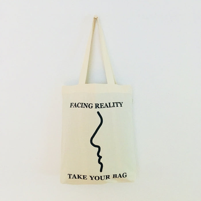 A beige cotton tote bag with a black text 'FACING REALITY' and 'TAKE YOUR BAG' design.