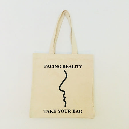 A beige cotton tote bag with a black design that reads 'FACING REALITY. TAKE YOUR BAG.'
