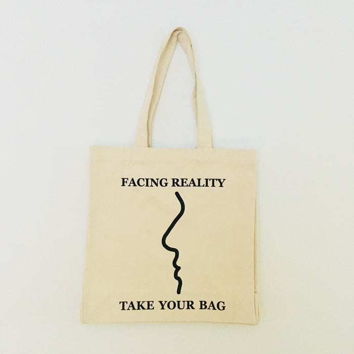 A beige cotton tote bag with a black design that reads 'FACING REALITY. TAKE YOUR BAG.'