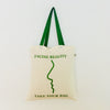 A beige canvas tote bag with a green text graphic design that reads 'FACING REALITY take your bag'.