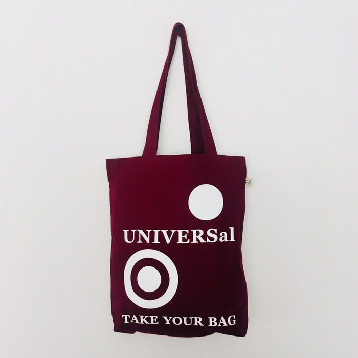 A burgundy tote bag with 'UNIVERSal' text and a graphic logo on the front, made of organic cotton, with a handle and a detachable strap.